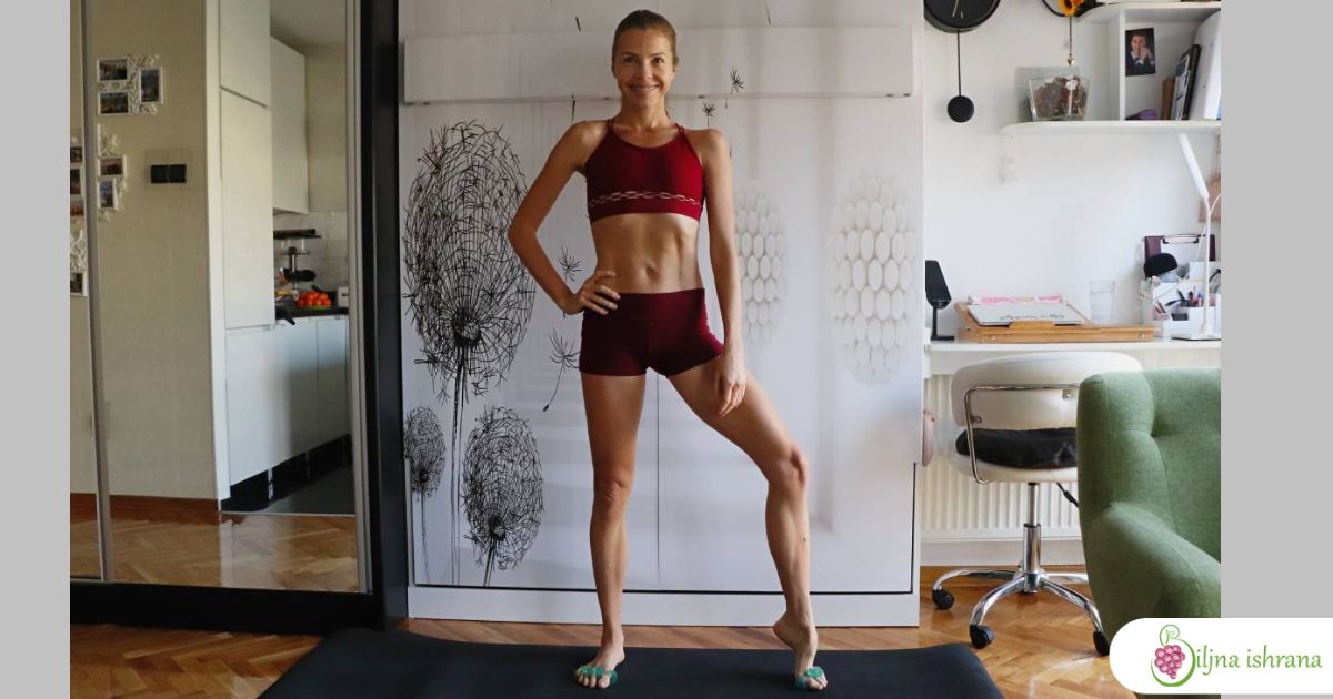 Marina sa Biljne ishrane stoji na prostirci za vežbanje u dnevnoj sobi. Na sebi ima crveni komplet za vežbanje.