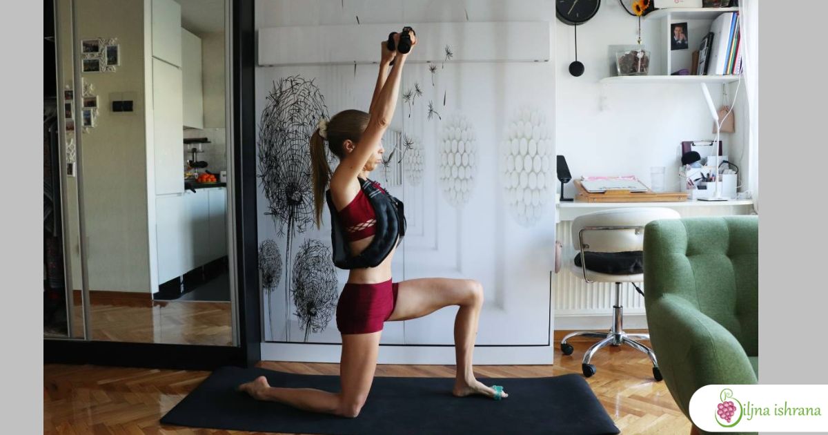 Marina sa Biljne ishrane kleči na jednoj nozi podignutih ruku u kojima drži tegove. Na sebi nosi prsluk sa opterećenjem.