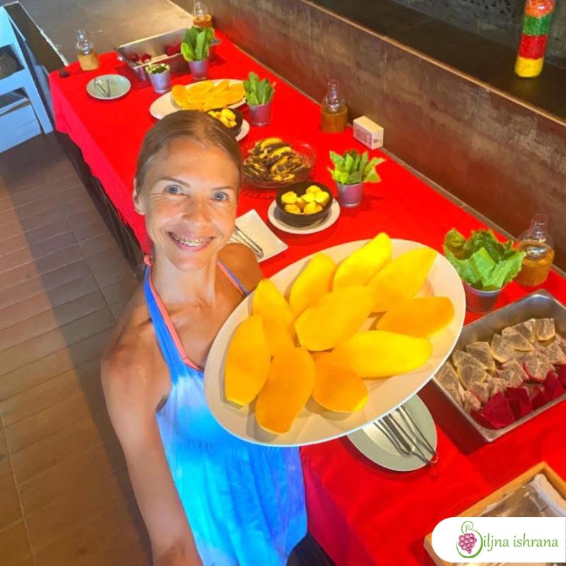 Marina sa Biljne ishrane drži tacnu punu manga i stoji pored švedskog stola na Tajlandskom ritritu Biljne ishrane.