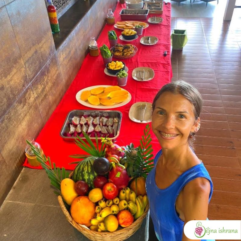 Marina sa Biljne ishrane stoji pored korpe pune tropskog voća. Iza nje je švedski sto na Tajlandskom ritritu Biljne ishrane.