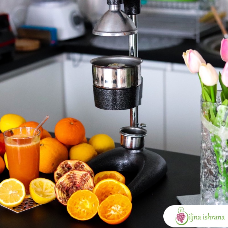 Presa za ceđenje nara slikana izbliza kako stoji na kuhinjskom pultu. Pored prese je citrusno voće i isceđene kore nara, limuna, narandže i grejpa.