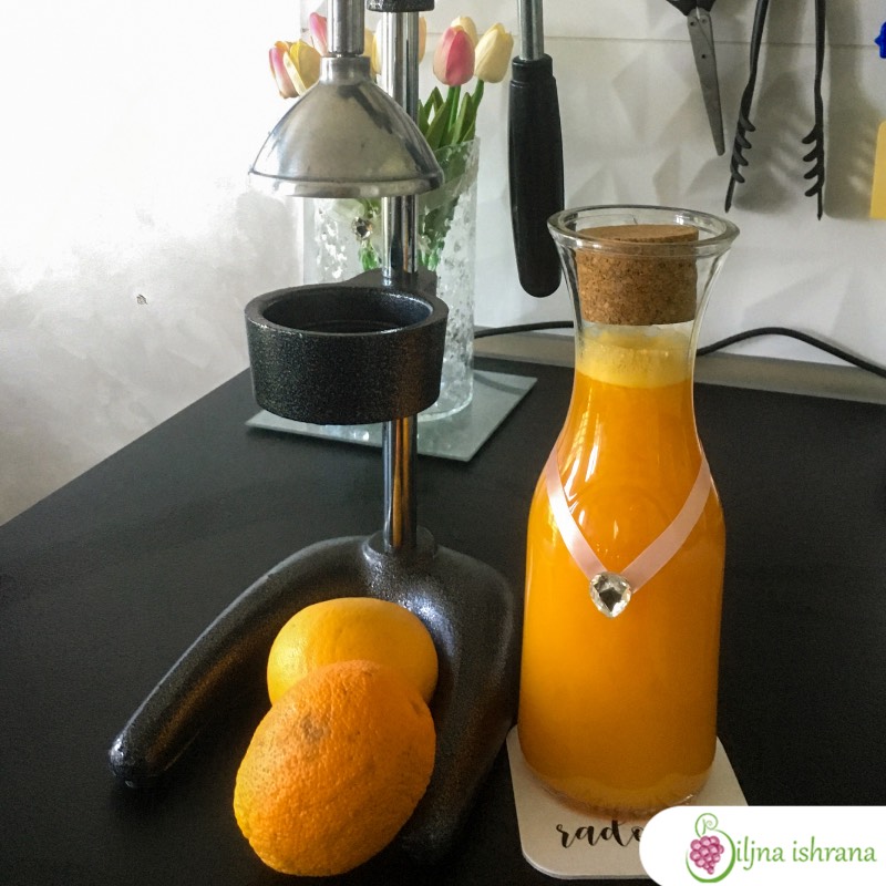 Presa za ceđenje nara stoji na kuhinjskom pultu. Pored prese je staklena flaša puna soka od narandže. Unutar osnove prese su dve narandže.