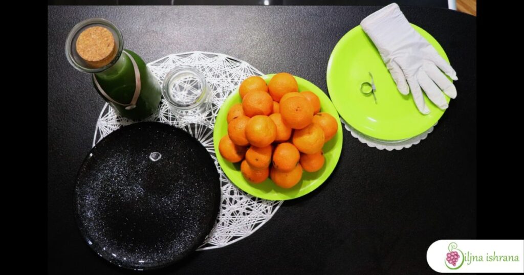 Sastojci i oprema za pripremu doručka na sirovoj ishrani zimi: flaša sa zelenim sokom, tanjir sa klementinama, tanjir za posluženje, tanjir za kore klementina, higijenska rukavica i guljač citrusa. Svi ovi elementi stoje na srebrnom podmetačnu i crnom stolu.