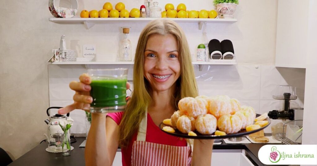 Marina sa Biljne ishrane drži tanjir pun oljuštenih klementina u jednoj ruci i čašu zelenog soka u drugoj ruci.