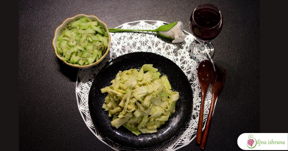 Tanjir sa sirovim veganskim makaronima sa sirom, drvena viljuška i kašika, činijica sa salatom od krastavaca i mikrobilja, vinska čaša sa sokom od nara, salveta u ukrasnom držaču, srebrni podmetač, crn sto.