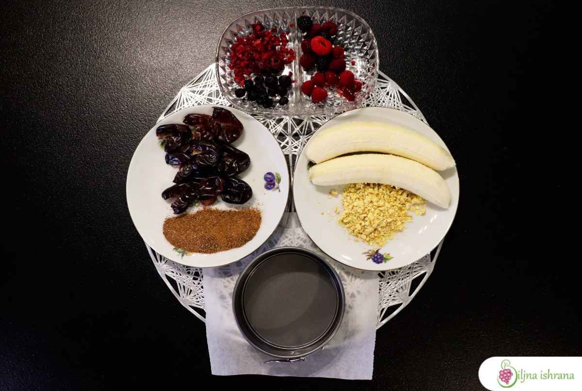 Sastojci za sirovu vegansku tortu i mali kalup za torte. Urme i rogač u tanjiru na levoj strani, bobičavo voće u staklenoj činiji na vrhu slike, banane i sušene jabuke na tanjiru na desnoj strani. Slikano od gore.
